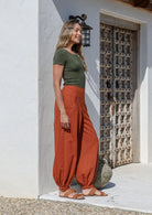 A woman wearing a green basic top and brick orange, genie style pull-on cotton pants with pockets, cuffed buttoned ankles, and a shirred elastic waistband.