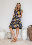 Woman wearing a cotton above knee dress with cap sleeves, pockets and generous skirt, poses in front of decorative vases.