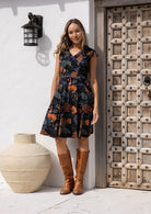Woman stands in sun wearing a beautiful above knee length cotton dress with leafy pattern in dark blue, green and grey with orange and pink florals on black base.