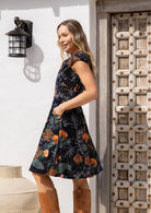 Model standing slightly sideways to the wooden door wears a gorgeous botanic print 100% cotton dress with leather boots and hands in pocket.