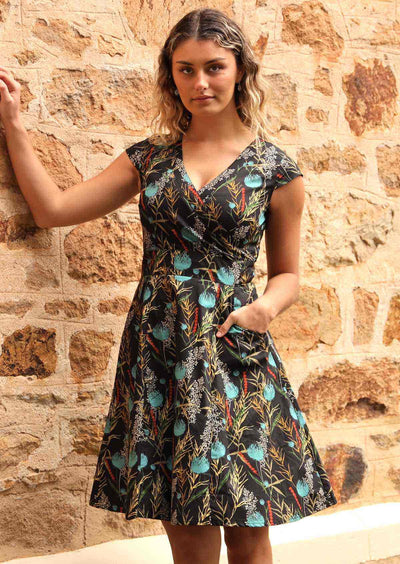 Model wears Alice Dress Thistle black base botanical print with teal highlights, 100% cotton, crossover bodice forming a V-neck, fuller skirt and pockets | Karma East Australia