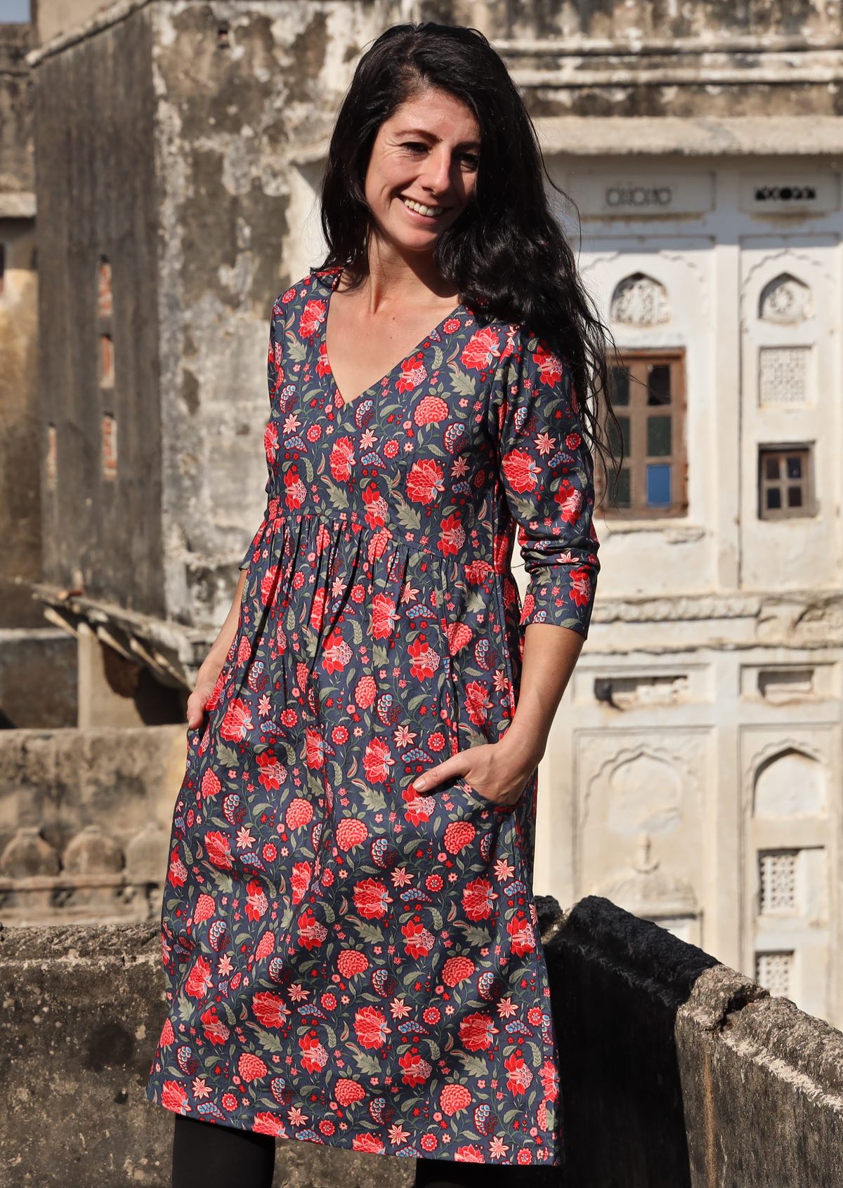 Woman wears cotton 3/4 sleeve V-neckline dress in a red and green floral print on a blue base, with hands in pockets