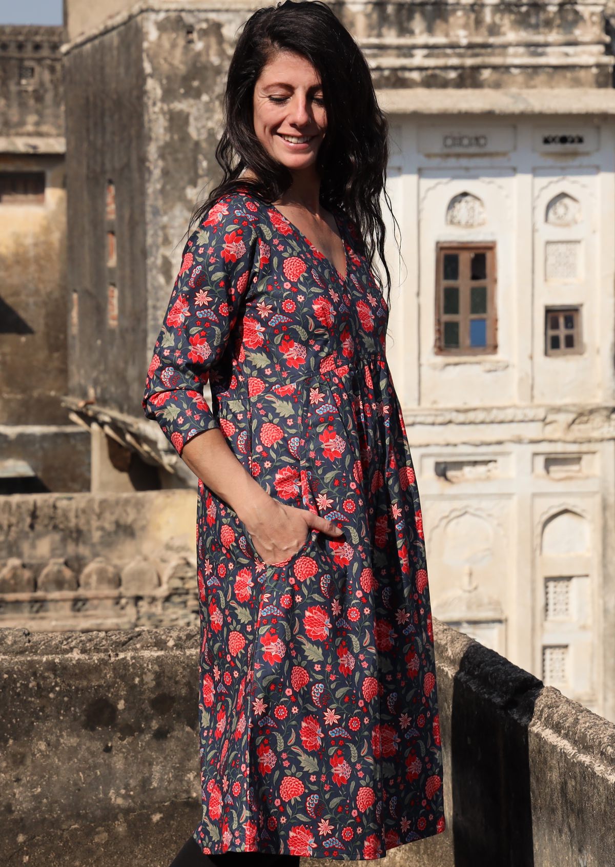 Woman stands side on in cotton 3/4 sleeve V-neckline dress in a red and green floral print on a blue base, with hands in pockets