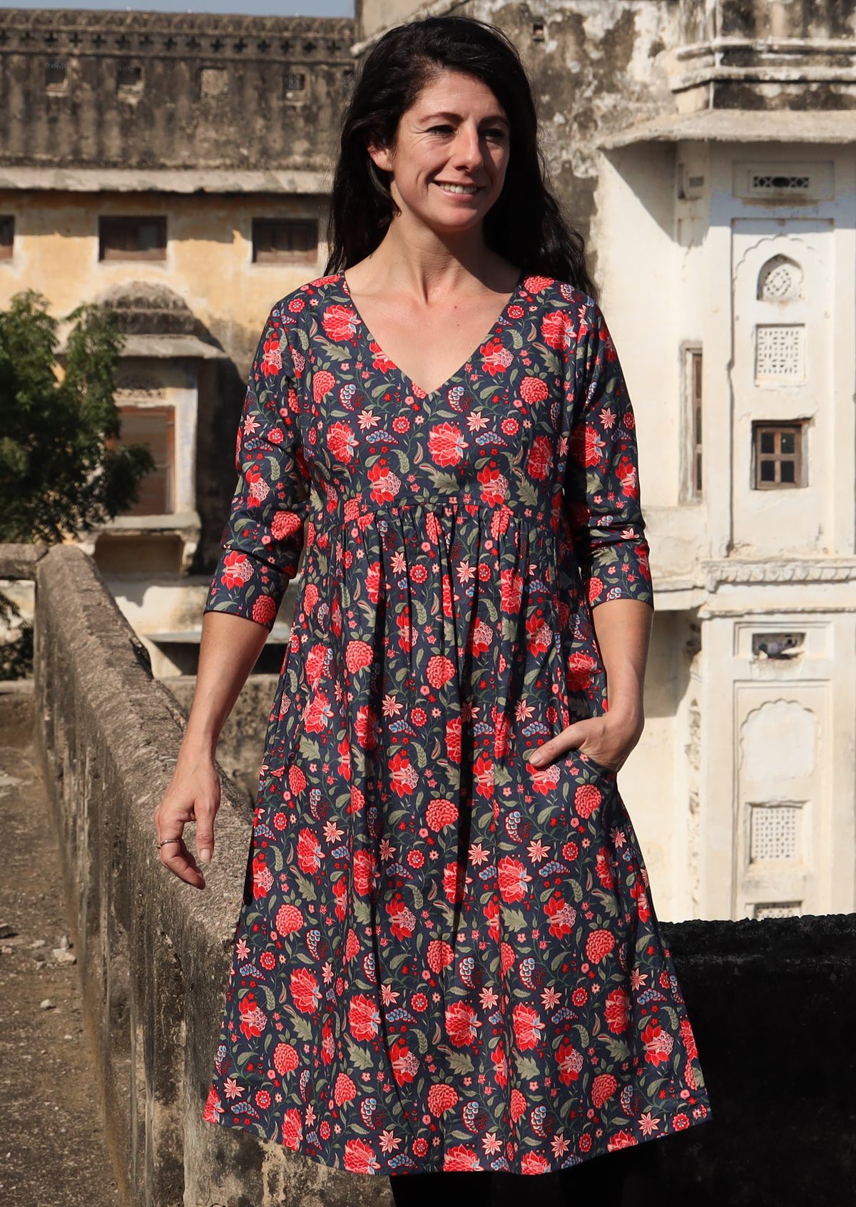 Woman wears cotton 3/4 sleeve V-neckline dress in a red and green floral print on a blue base