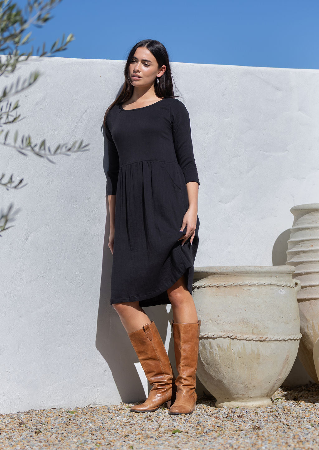 Woman in a black cotton dress and brown boots standing against a white wall with a blue sky.