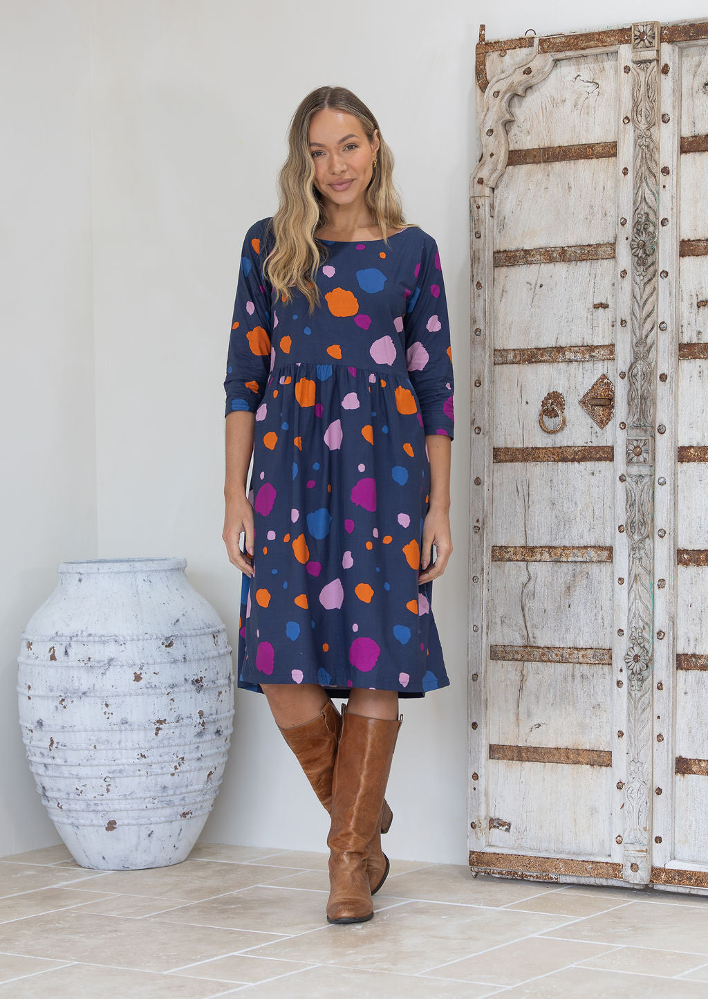 Woman wearing a blue based dress with colourful polka dots standing next to a wooden door.