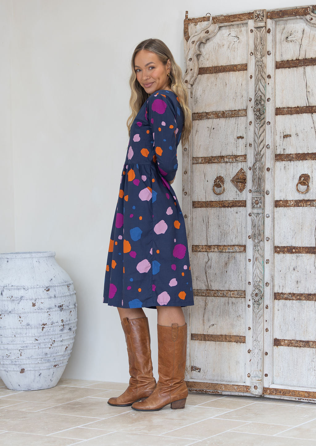 Woman stands side on in a navy blue base dress with colourful polka dots and tan knee-high boots