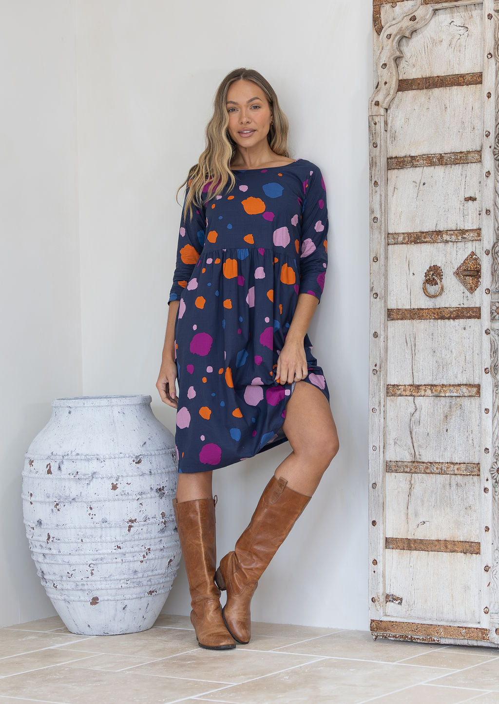 Woman wearing a cotton knee length dress with orange, blue, plum and pale pink spots on a navy blue base, paired with tan boots
