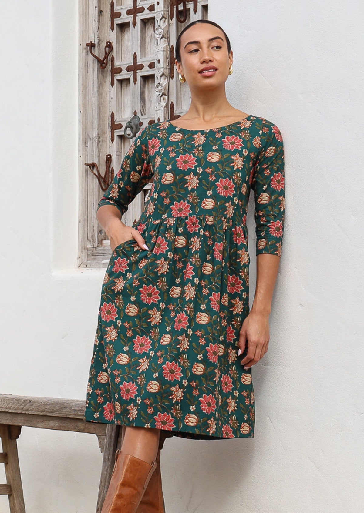 Woman in deep green, floral print cotton,  knee length dress with 3/4 sleeves, wide round neckline and a hand in pocket.