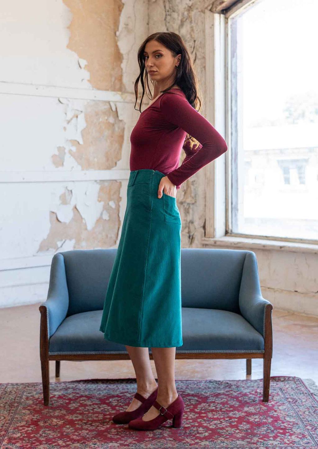 Woman stands side on in teal corduroy A-line midi length skirt with hands in the back pockets