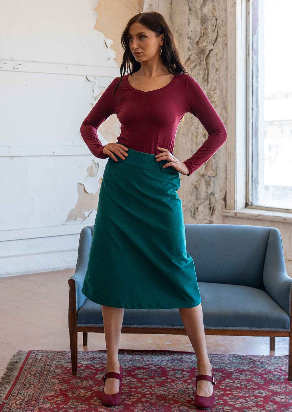 Model poses in teal corduroy midi length skirt with belt loops, paired with a maroon long sleeve rayon top