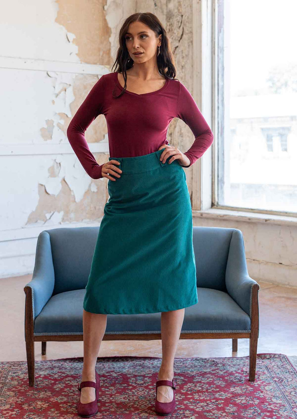 Woman wears corduroy A-line midi length skirt in teal, paired with a long sleeve maroon top