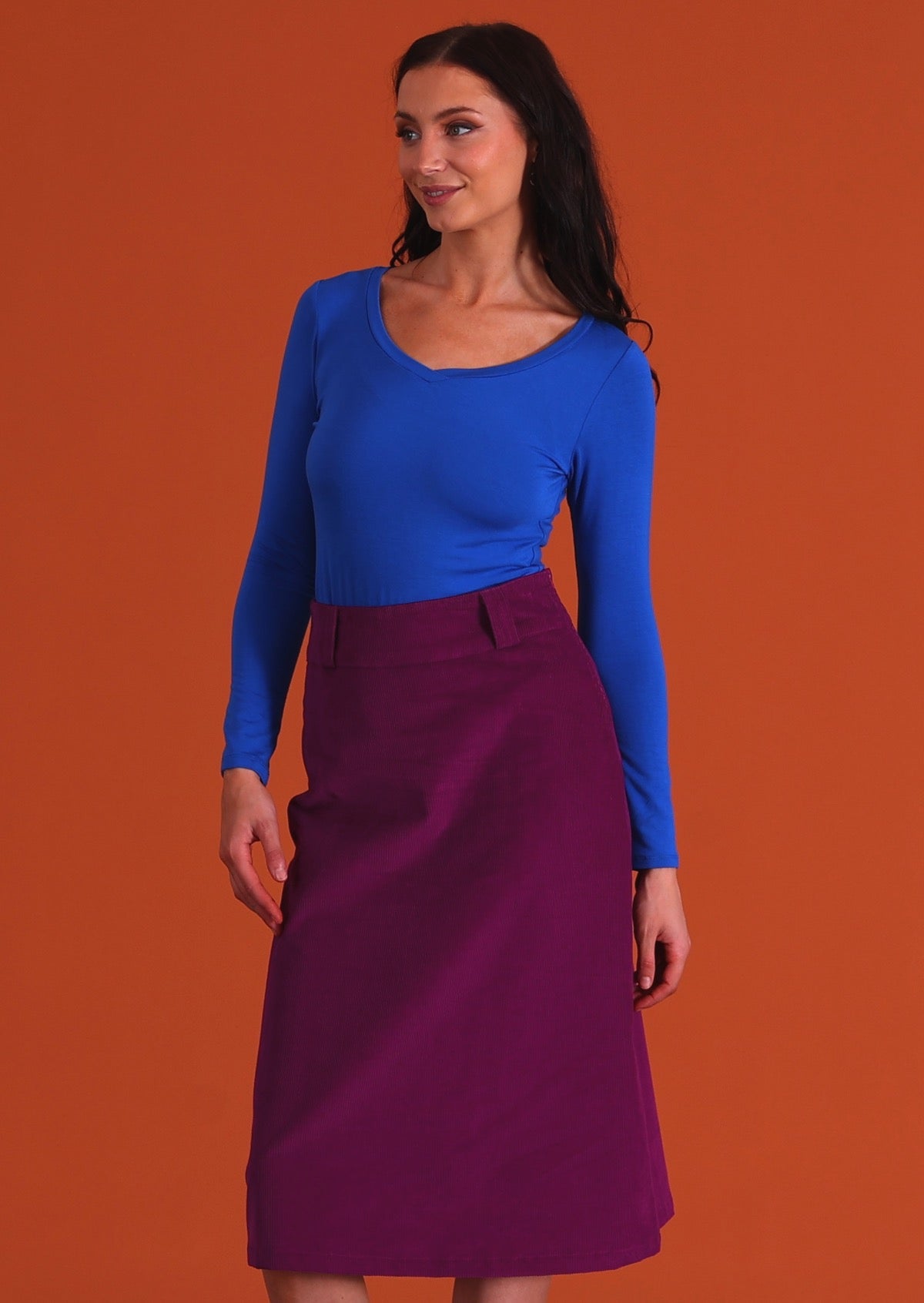 woman wearing purple corduroy aline skirt  with belt loops with bright blue long sleeve top with orange back drop
