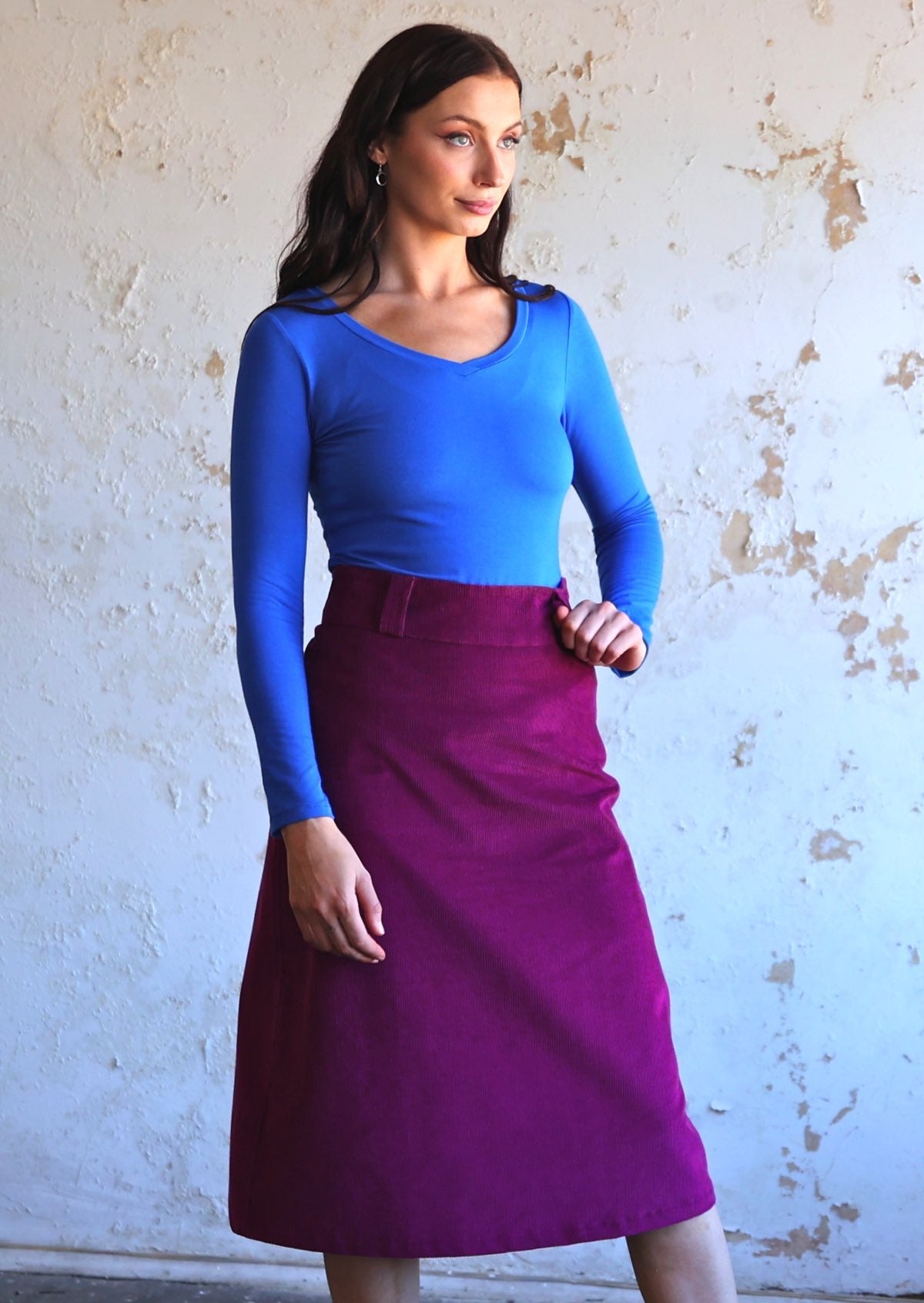 woman wearing purple corduroy aline skirt  with belt loops with bright blue long sleeve top with v-neckline