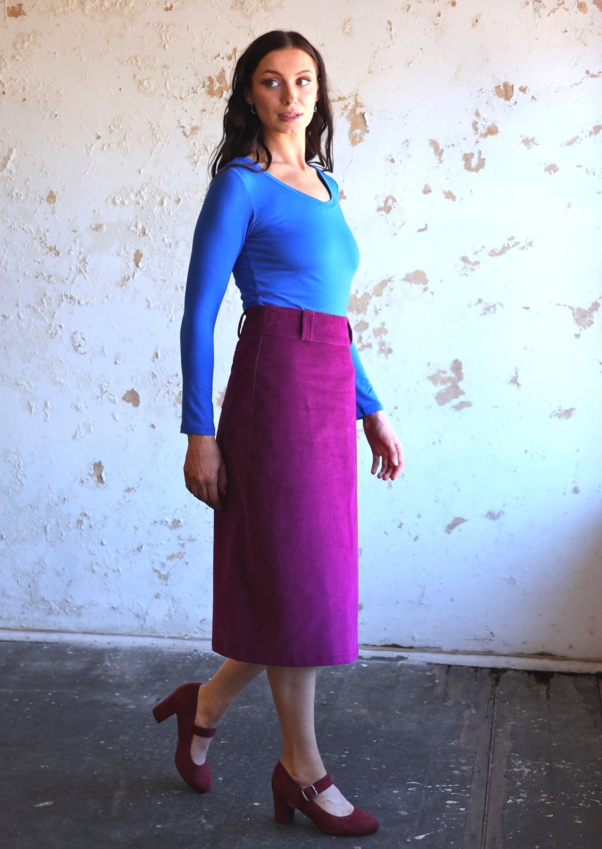 woman wearing purple corduroy below the knee aline skirt  with belt loops with bright blue long sleeve top