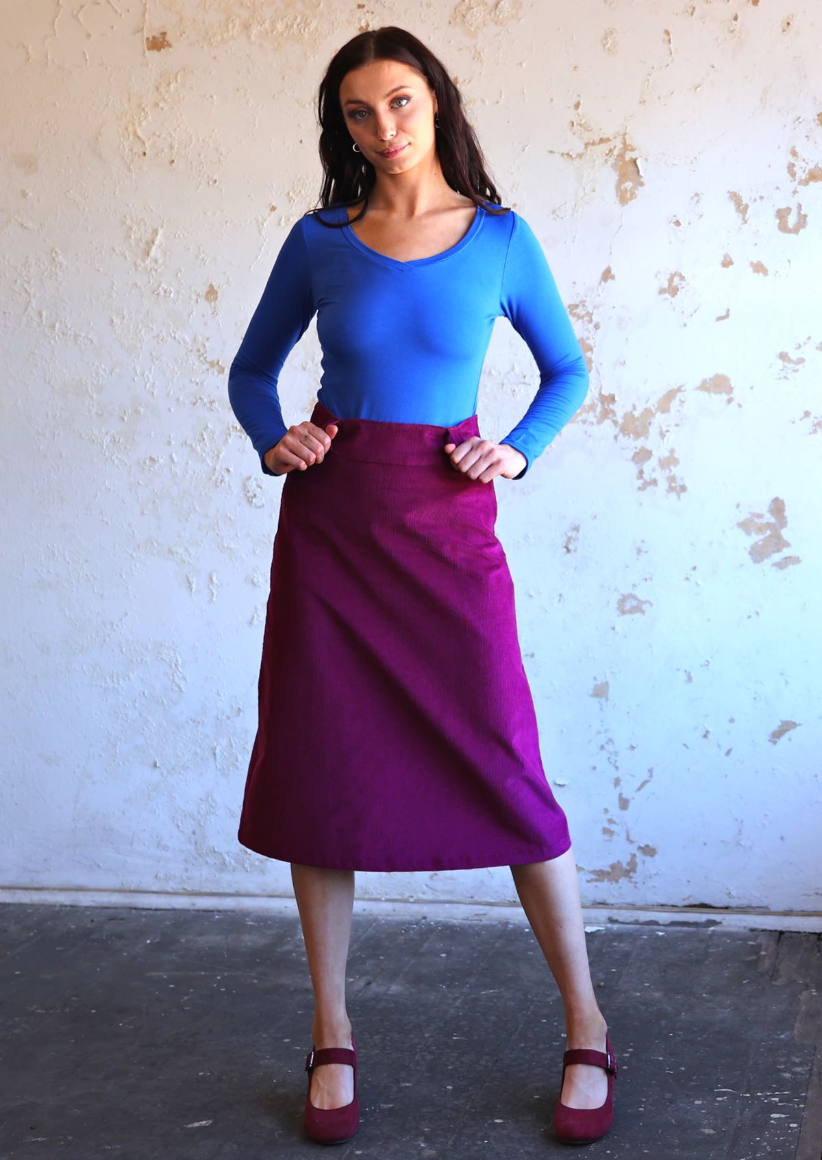 woman wearing purple corduroy aline skirt  with thumbs through belt loops with bright blue long sleeve top