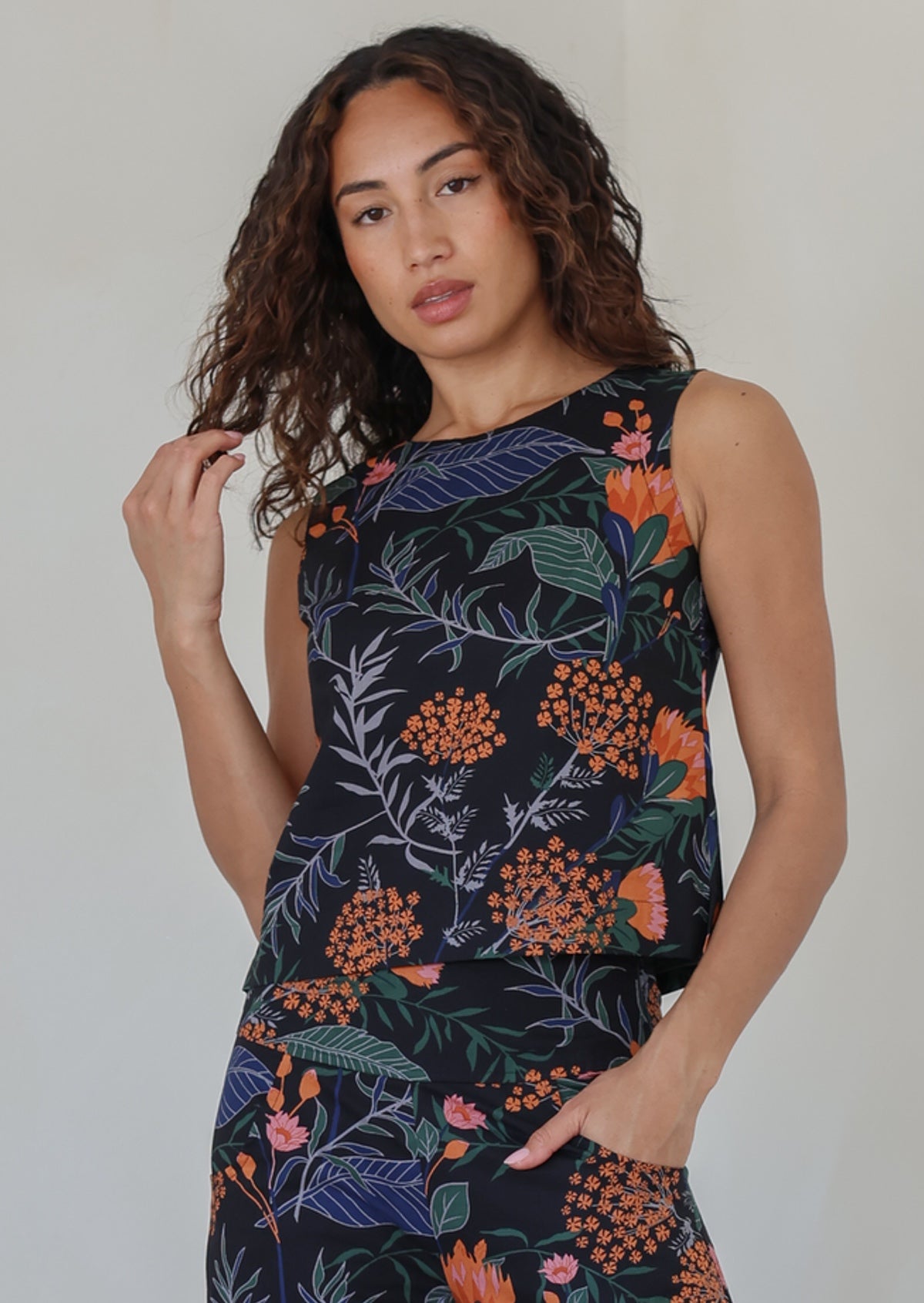Woman with dark hairs posing in a sleeveless black based botanical print top featuring high round neckline 