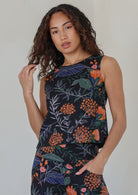 Woman with dark hairs posing in a sleeveless black based botanical print top featuring high round neckline 