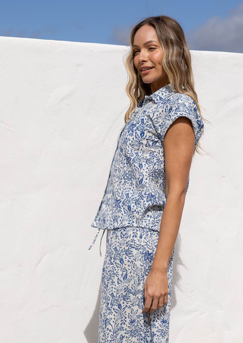 Woman wearing a white based blue floral print cotton shirt which has short sleeves against a white background.