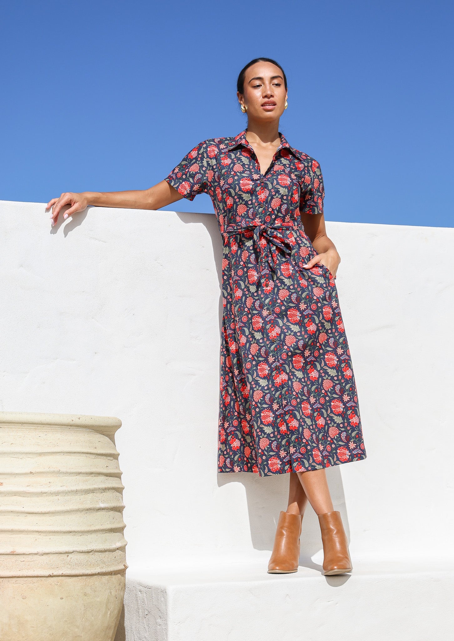 Woman poses in cotton navy blue based floral print shirt dress with fabric belt, and hand in pocket 