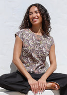 Woman sitting on a white surface wearing printed cap sleeved cotton top in grey with delicate beige, pink and lime floral print 