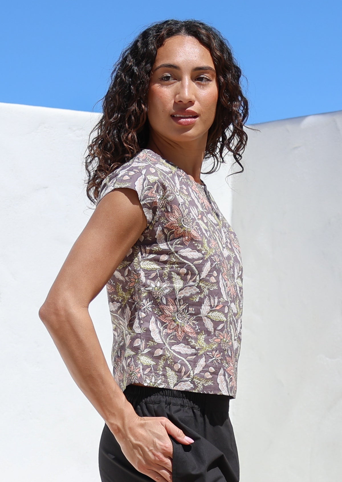 Woman stands in sun with hands in pants pocket wearing a 100% cotton floral print top featuring high round neckline and cap sleeves 