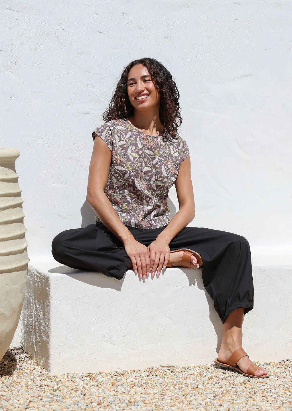 Woman sitting on a white concrete bench wears grey based floral print top featuring round neckline and cap sleeves 