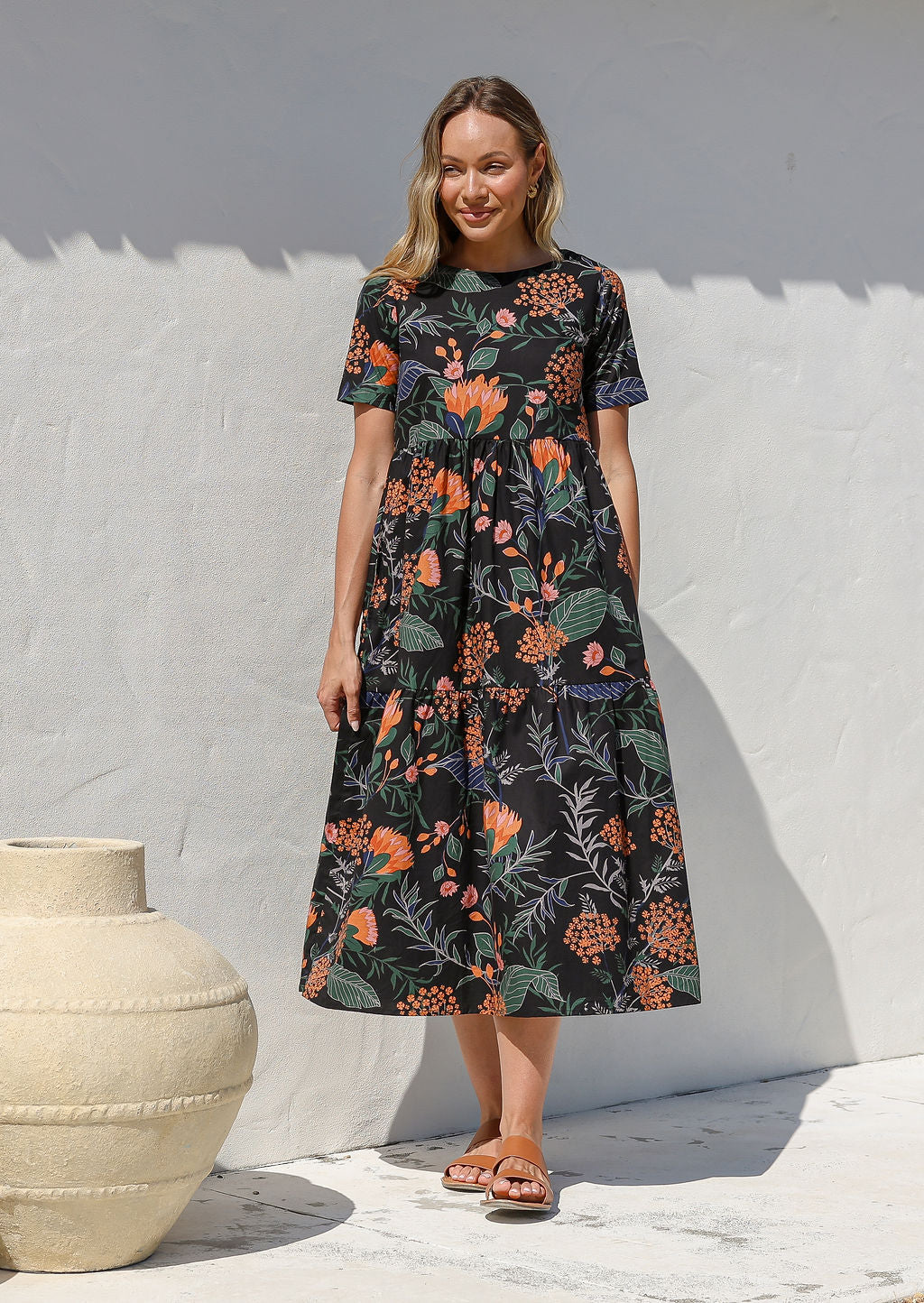Woman standing against the white wall wears a floral black cotton dress with three tiered design, short sleeves and pockets