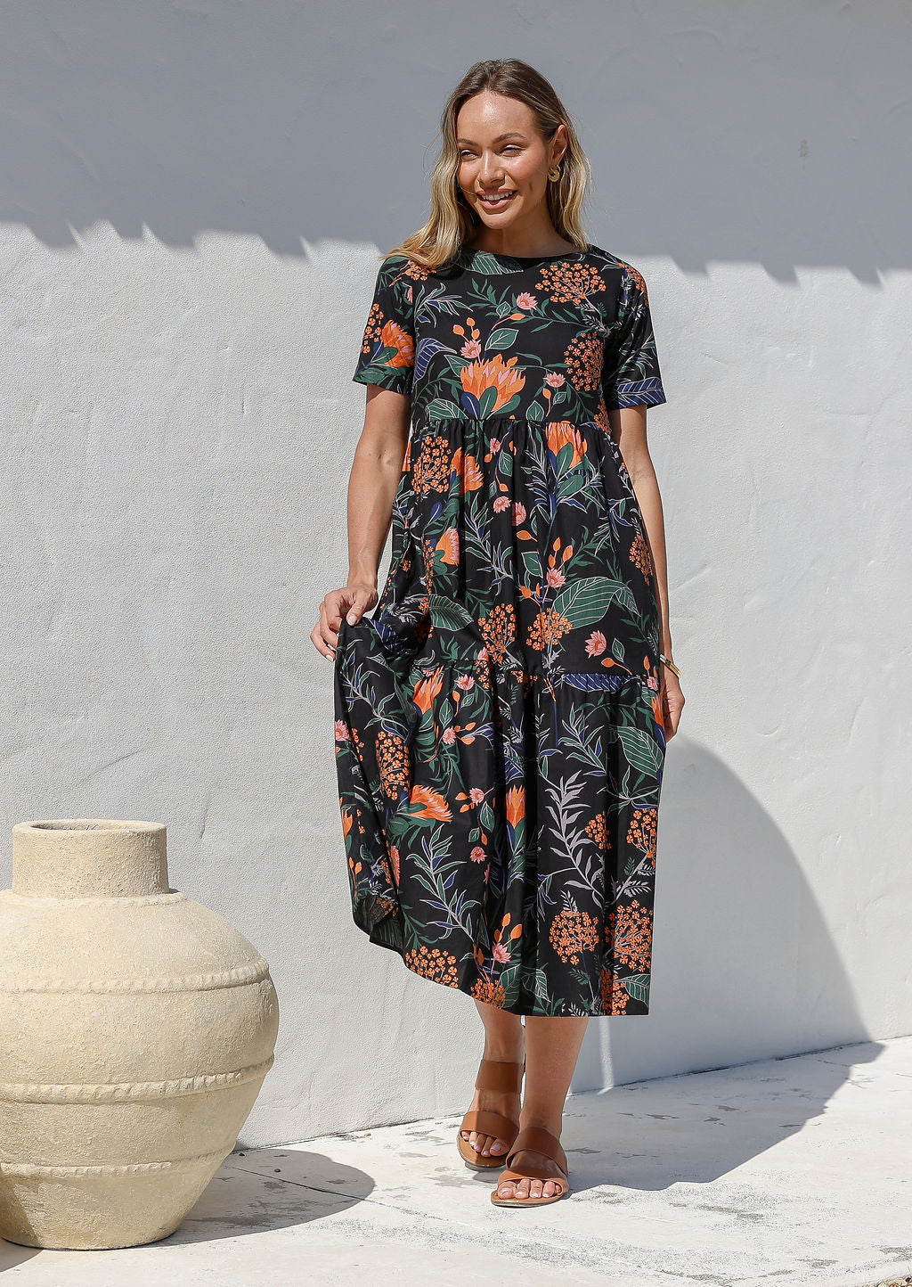 Woman is wearing a relaxed fit midi length, tiered cotton dress in black based botanical print with short sleeves, round neckline and side pockets.
