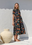 Woman wearing a relaxed fit printed black cotton dress with tiered design, standing next to a large vase against a white wall