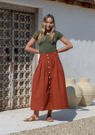 Woman wearing a green basic top and rust colored shin length, A line cotton skirt with box pleats, belt loops and button through front