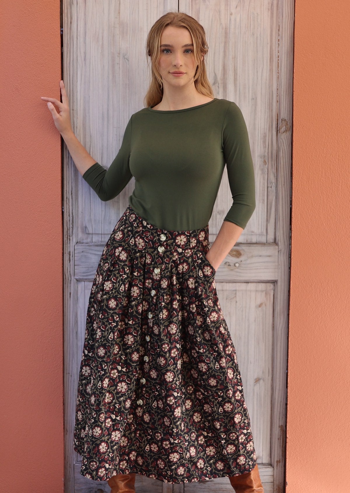 Woman standing in front of white door with one hand on door frame wears black based white and maroon floral print cotton button through skirt other hand in pocket