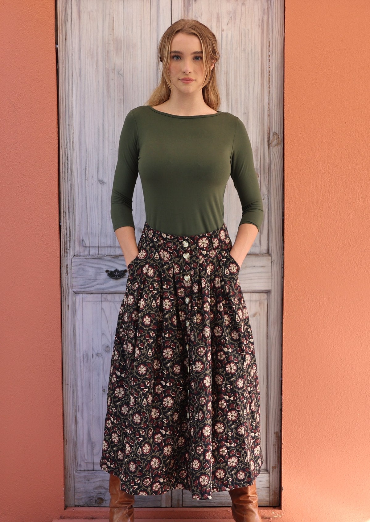 Woman standing in front of white door wears black based white and maroon floral print cotton button through skirt with hands in pockets