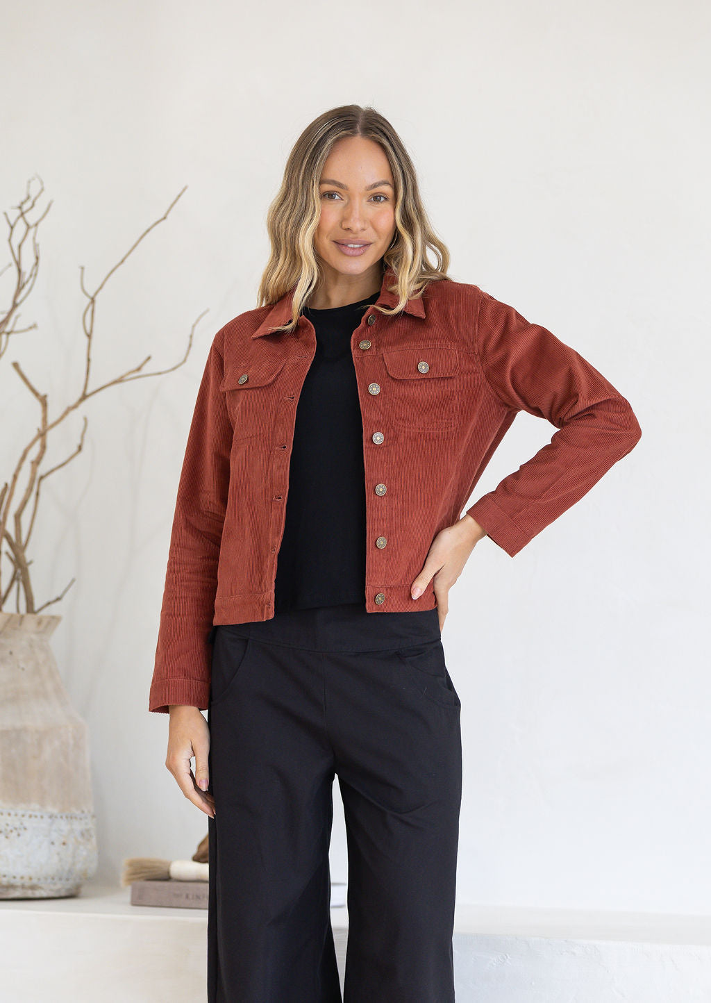 Woman wearing a rust-colored jacket over a black top and pants, standing against a light background.