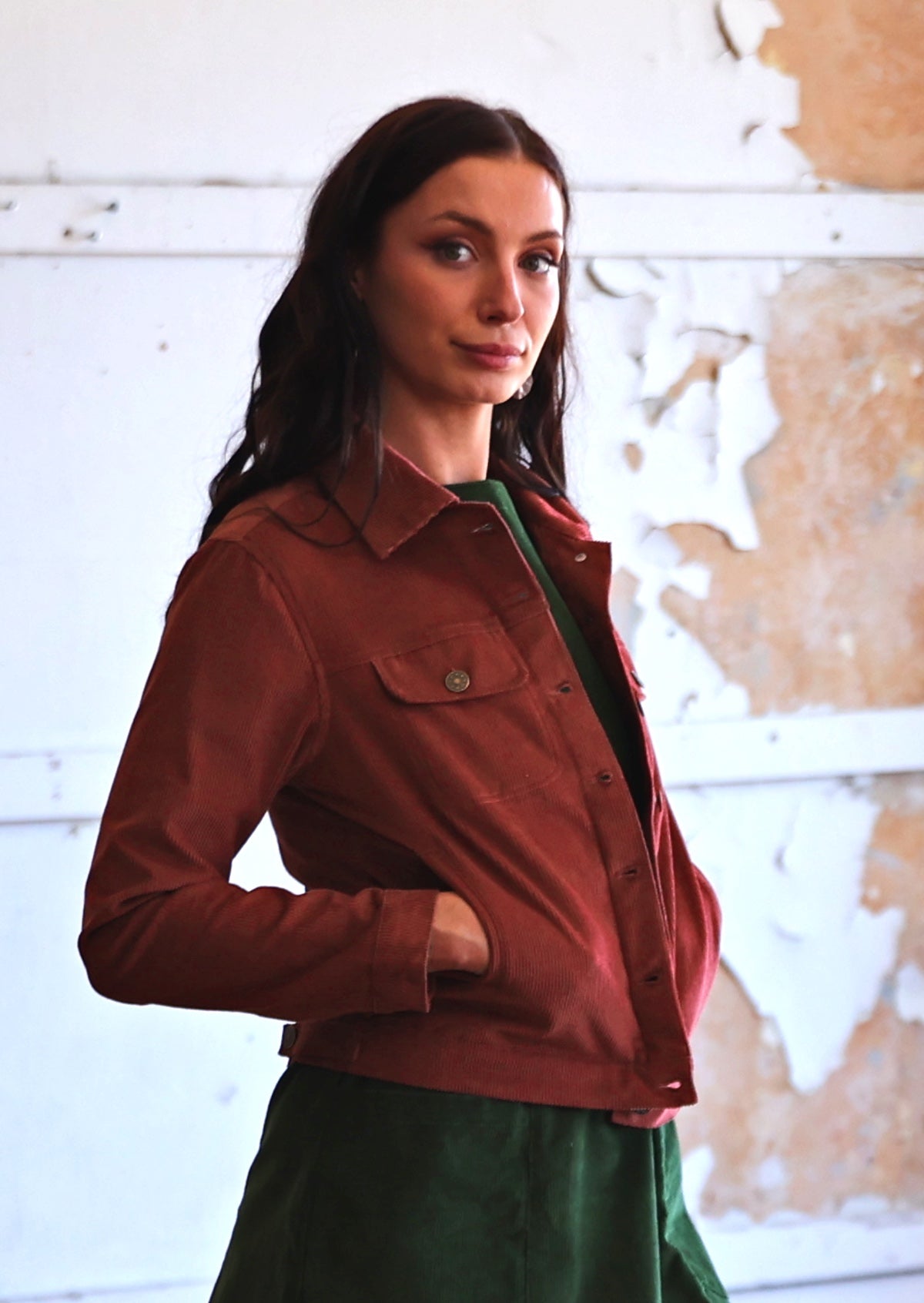 Woman in rust coloured cotton corduroy jacket over green corduroy dress with hands in pockets 