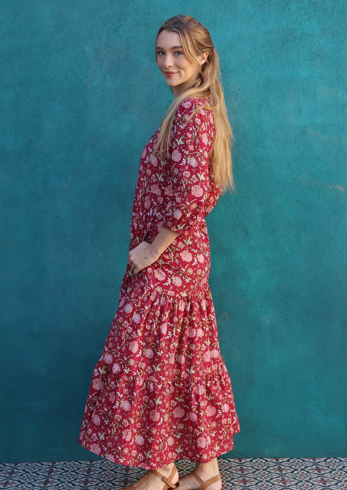 Woman stands sideways in cotton tiered maxi dress in beautiful bright pinky red floral print, with thumb hooked in pocket