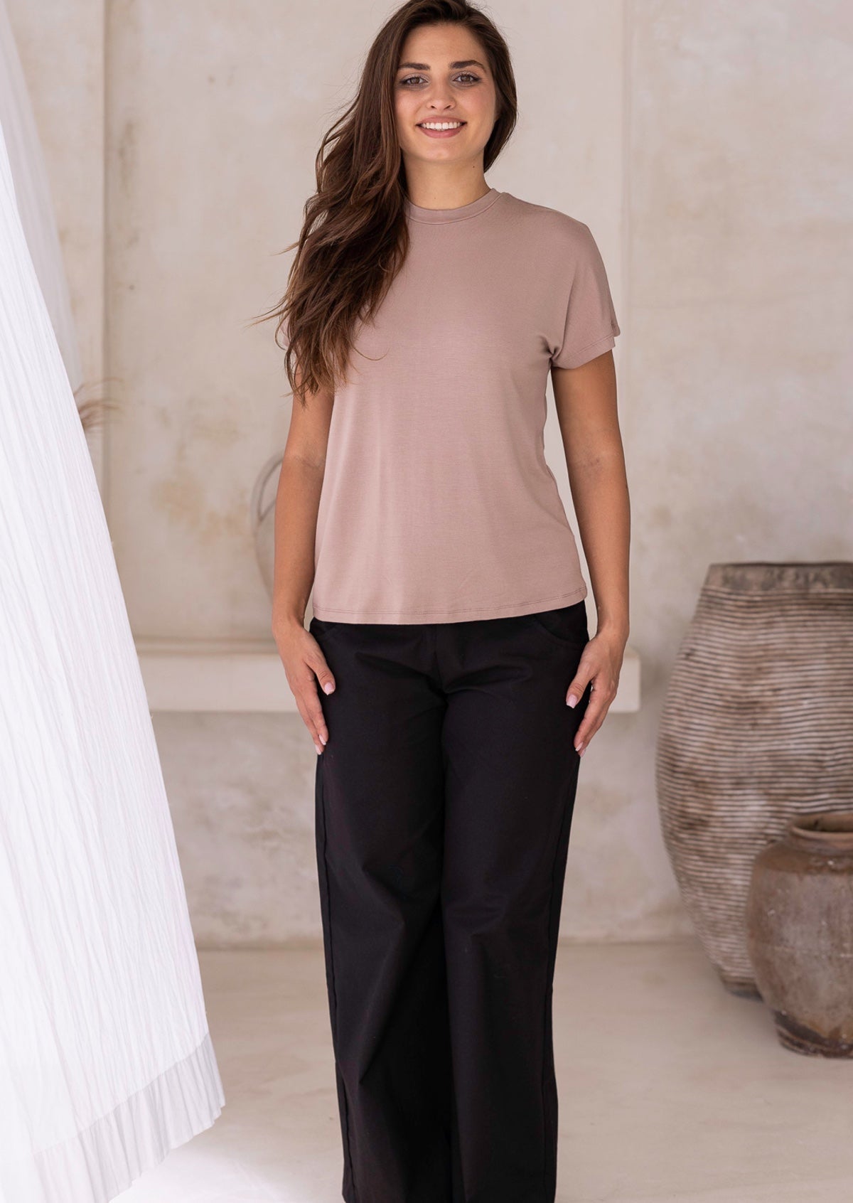 Woman wearing a beige t-shirt and black pants standing in a neutral setting with a vase in the background.