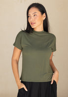 Woman wearing an olive green t-shirt against a beige background, with hands in pockets
