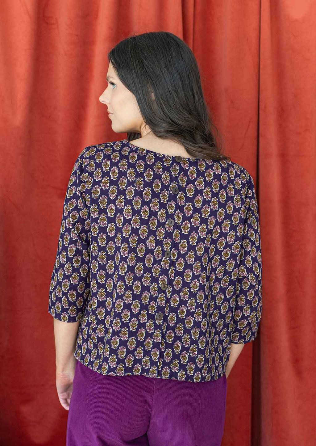 Model stands with her back turned wearing a relaxed fit cotton top with decorative buttons down at the back.