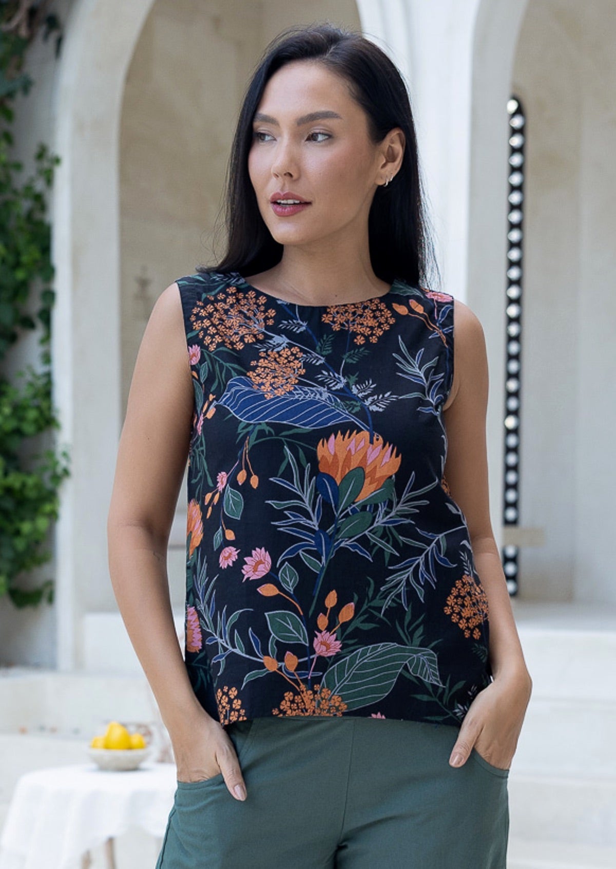 Model standing with hands in pocket of pants, wearing a sleeveless cotton top with round neckline and A line cut in beautiful bold botanical print on black base 