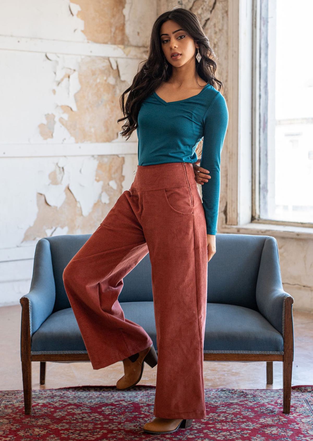 Woman wears burnt henna coloured corduroy wide legged pants with a side zip and pockets, paired with a teal long sleeve rayon top