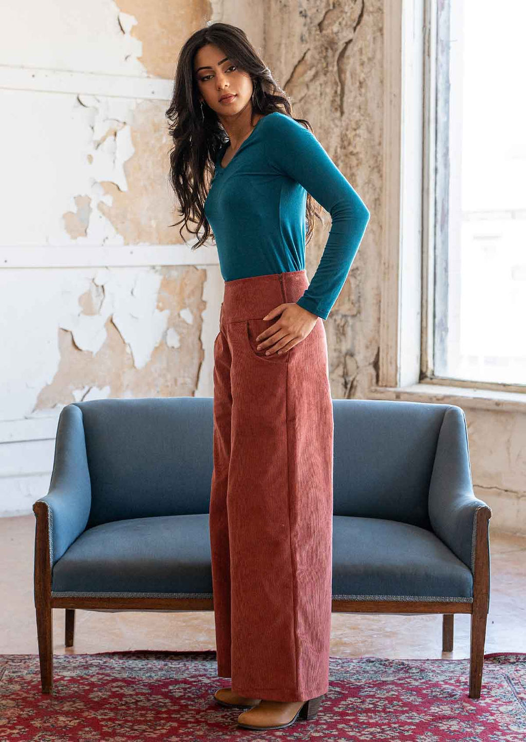 Woman stands side on in terracotta coloured wide leg corduroy pants with wide waistband, paired with a teal long sleeve top