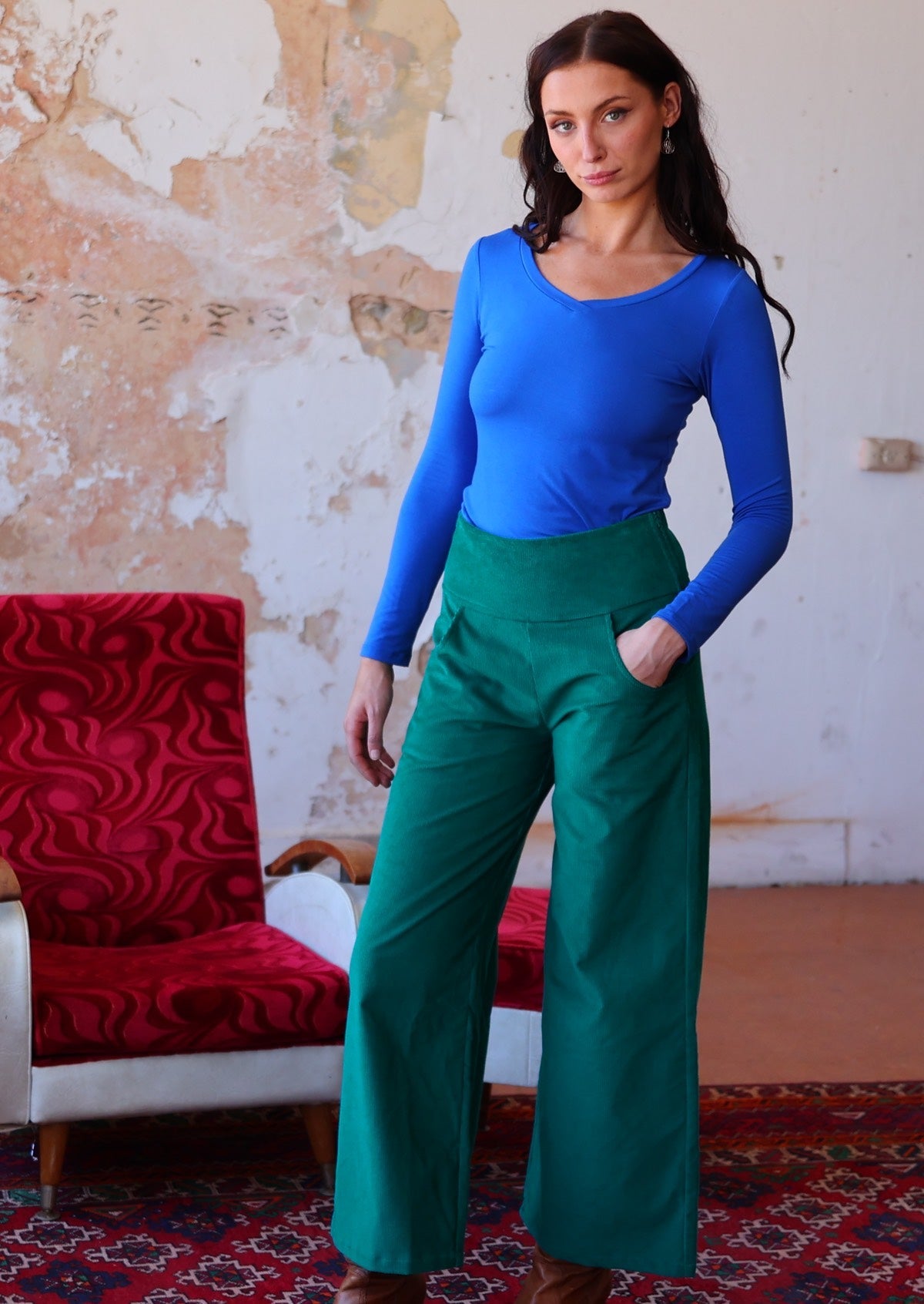 Woman wears electric blue long sleeve top with teal wide leg corduroy pants with a wide waistband, with her hand in pocket