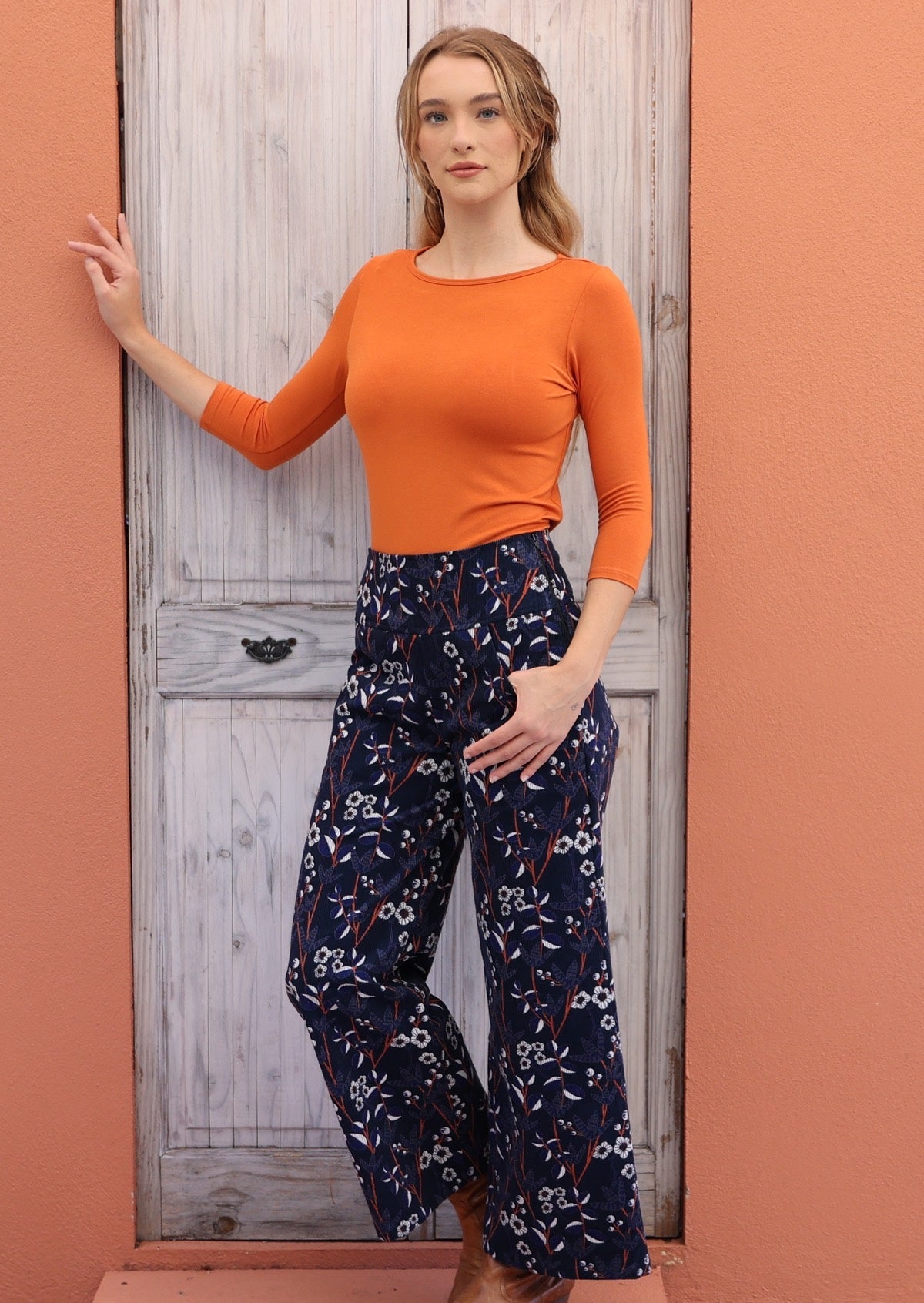 Model wears blue, orange and white floral print on dark blue base cotton corduroy pants with one hand in pocket.