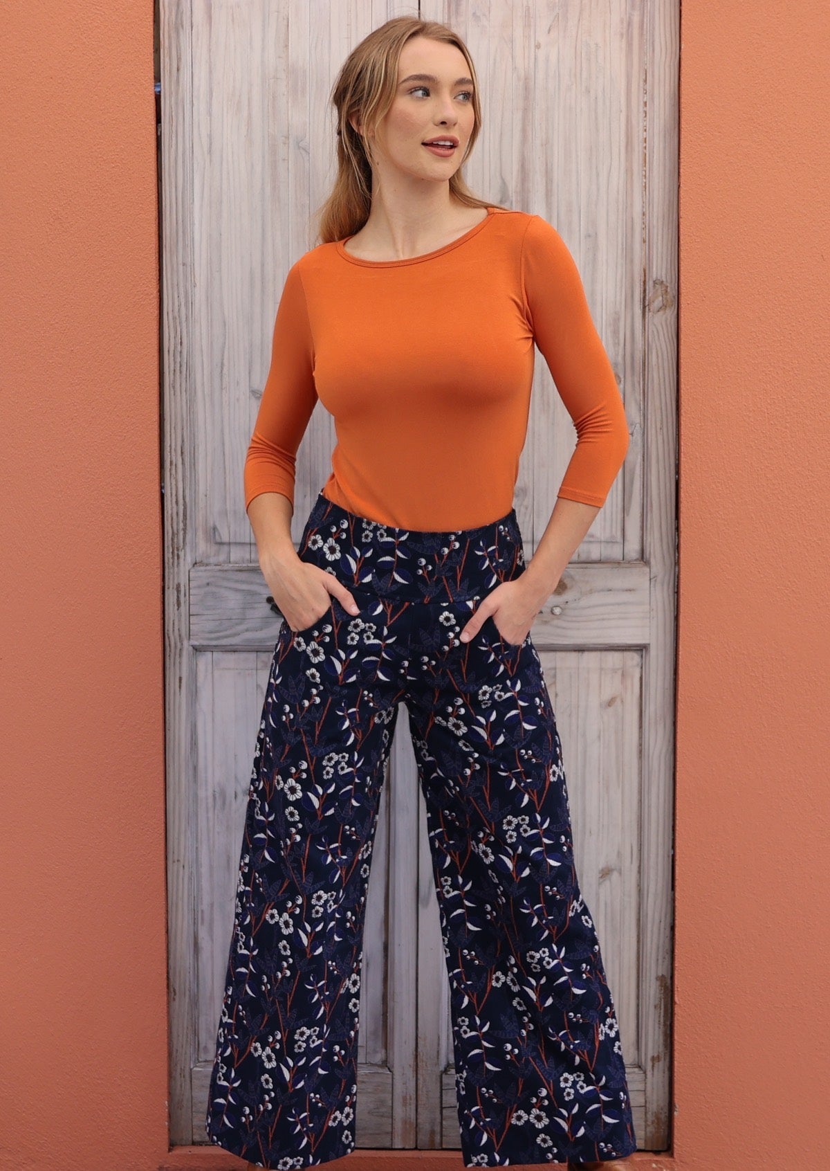 Woman wears blue, white and orange  floral print on navy blue base cotton corduroy pants with hands in pockets.