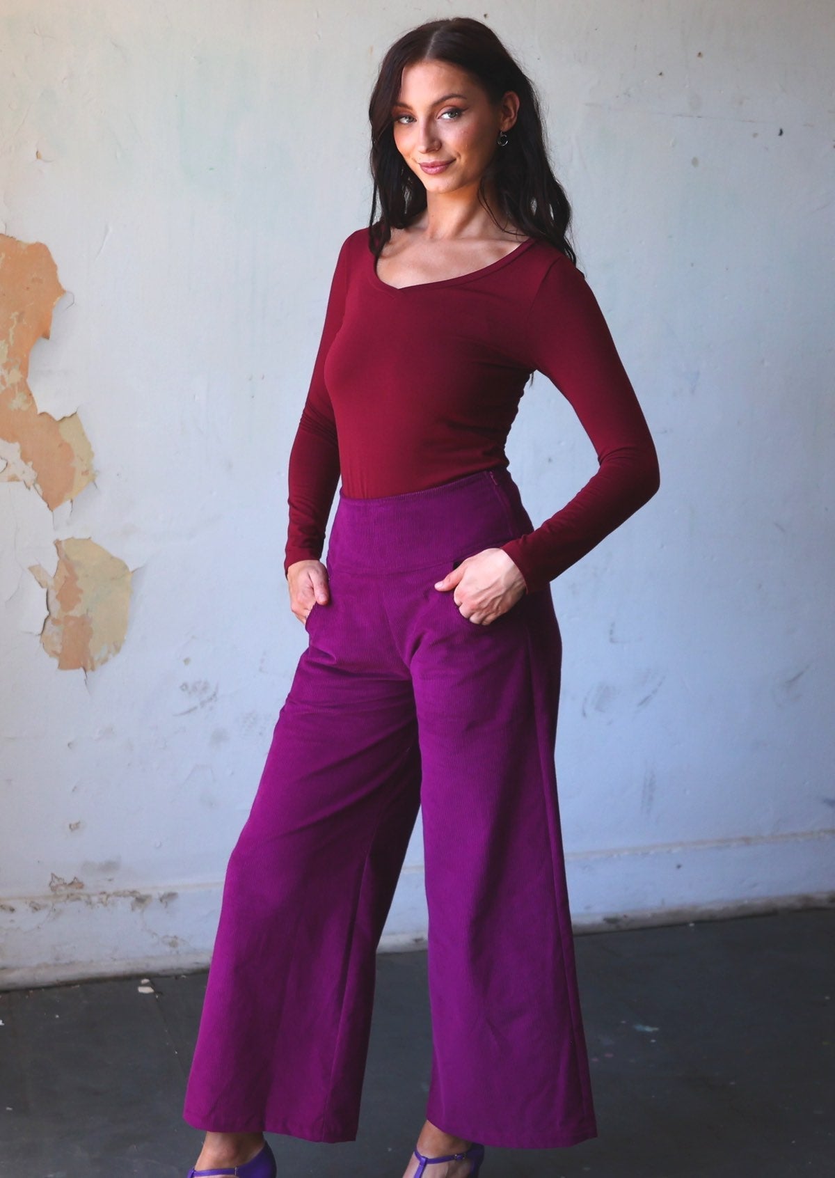 Woman poses in plum purple wide leg corduroy pants with hands in pockets, paired with a maroon long sleeve stretch rayon top