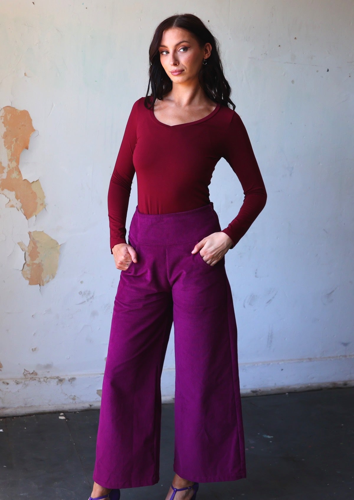 Model wears plum purple corduroy wide leg pants with her hands in pockets, paired with a maroon long sleeve V-neck stretch rayon top