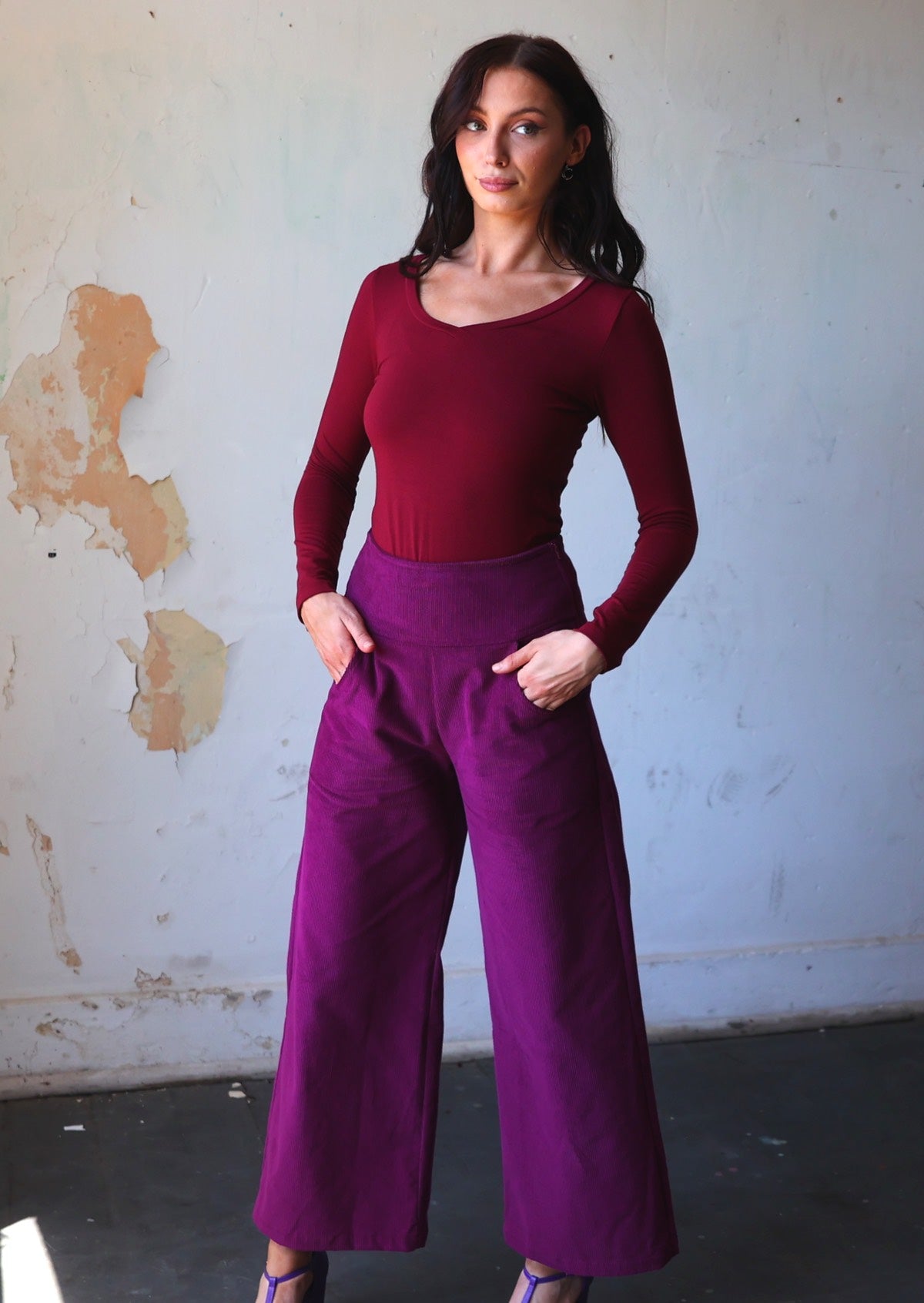 Model wears plum purple corduroy wide leg pants with a wide waistband and side zip, with her hands in pockets