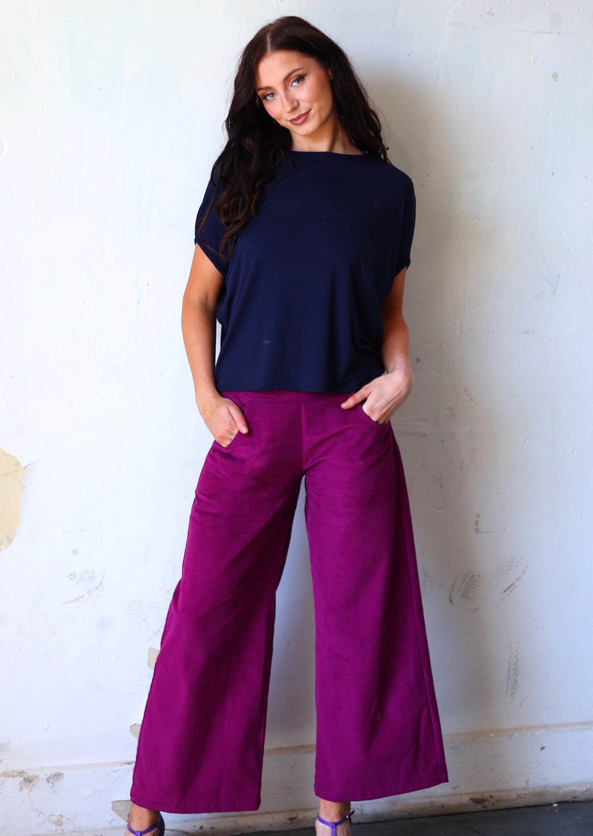 Woman poses in plum purple wide leg corduroy pants with hands in pockets, paired with a dark navy batwing top