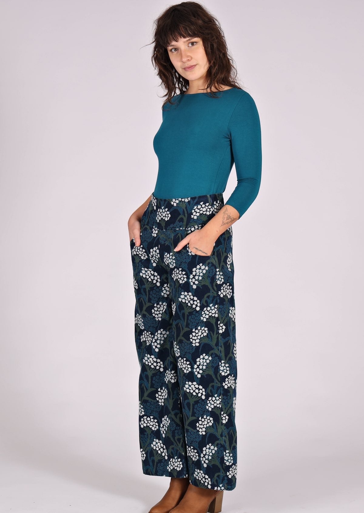 Woman stands in teal top and wide leg corduroy pants in floral print on a dark blue base
